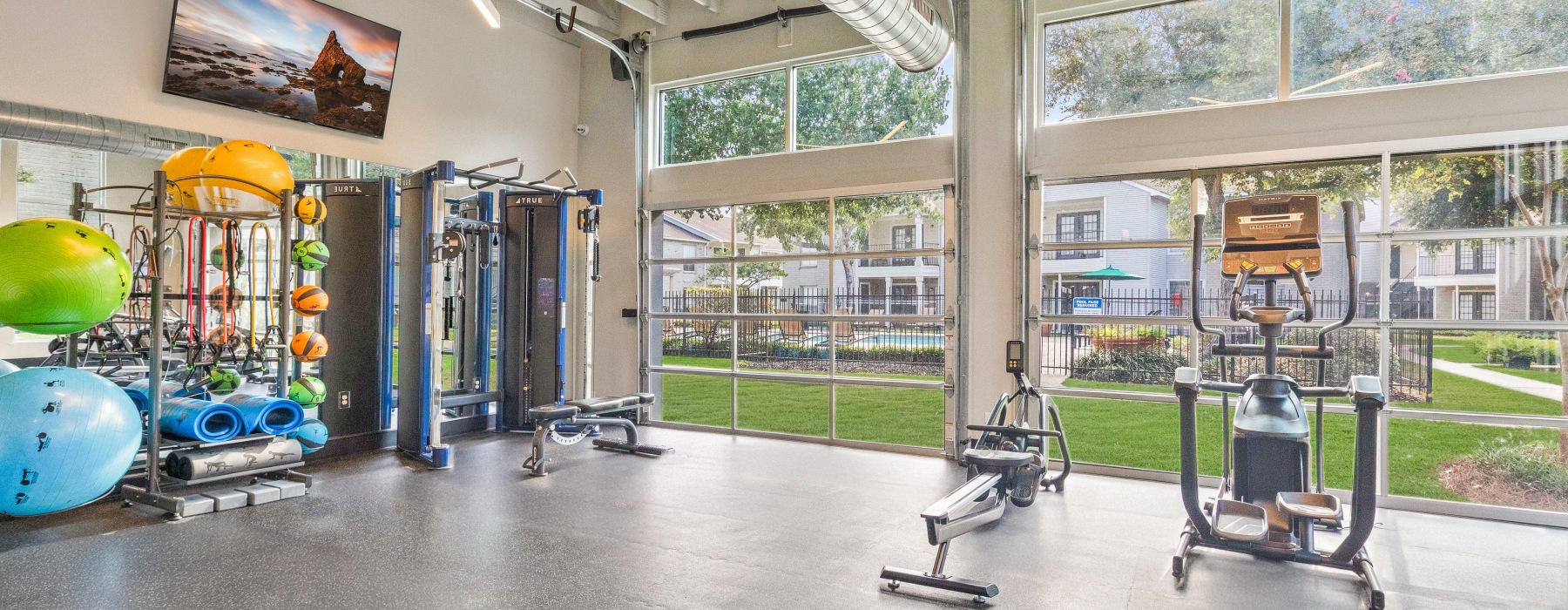 Spacious apartment gym featuring free weights, resistance machines, and bright natural light near the pool area in East Baton Rouge.